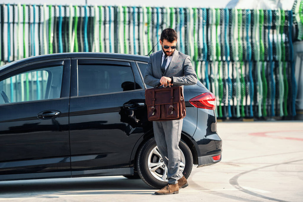 Full length of attractive caucasian rich businessman standing next to his expensive car, holding briefcase and looking at wristwatch. He don't want to be late for meeting. - Фото, зображення