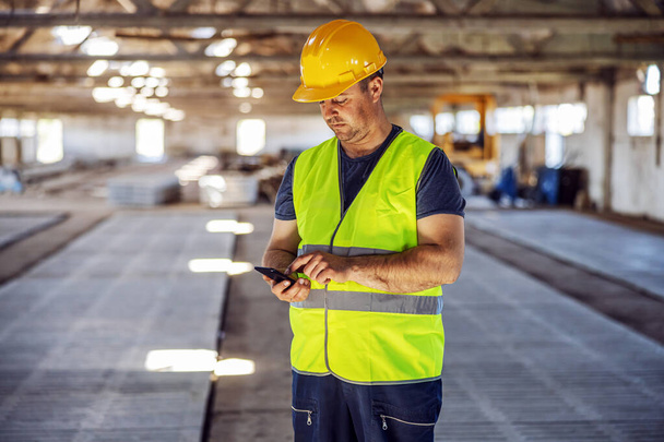 Lavoratore operaio blu in piedi in cantiere e utilizzando lo smartphone per ordinare altri materiali per la costruzione
. - Foto, immagini