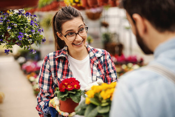 Mladá smějící se zahradnice stojí se svým kolegou a drží květináč s červenými květy. Selektivní zaměření na ženu. - Fotografie, Obrázek