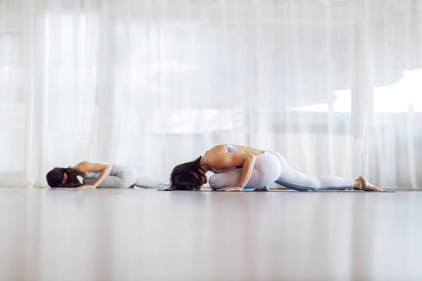 Twee aantrekkelijke fit yogi meisjes in Sleeping Swan yoga pose. Yoga studio interieur. - Foto, afbeelding