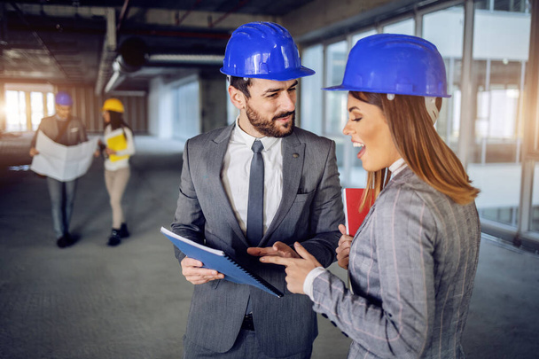 建設プロセスで建物の中に立っている頭の上にヘルメットと2人の笑顔魅力的な建築家は、書類を保持し、建物の更新のための新鮮なアイデアについて話しています。工事現場 - 写真・画像