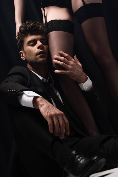 enfoque selectivo de hombre guapo en traje tocando pierna de mujer sexy en medias y sentado en negro
   - Foto, imagen