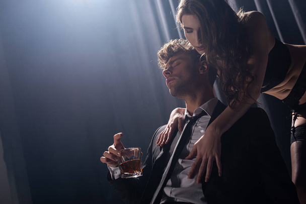 svůdná dívka dotýkající se pohledný muž v obleku sedí a drží sklenici whisky na černé - Fotografie, Obrázek