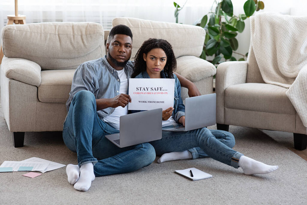 Serious african couple sitting with laptops on floor and holding warning poster - Photo, Image