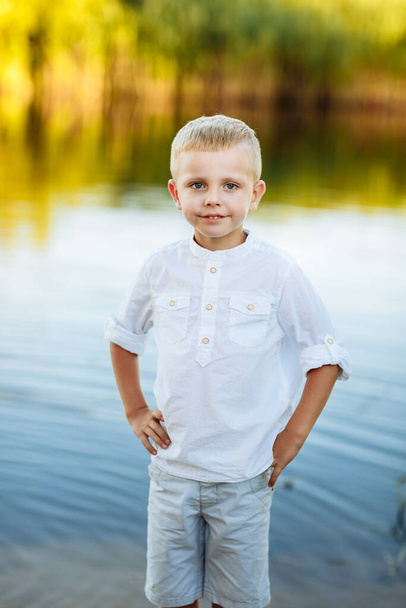 ブロンドの髪と白いシャツ、幸せな子供時代を持つ幸せで美しい小さな男の子の肖像画。肯定的な感情。自然界の川の近くの肖像画. - 写真・画像