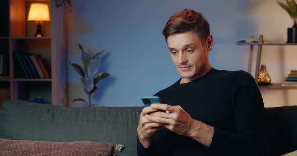 Young handsome man eading shocking news while using his smartphone . Cheerful guy in 30s making amazed face and big eyes while looking at phone screen and sitting on sofa at home. - Materiaali, video