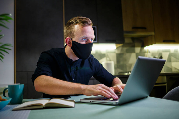 Νεαρός επιχειρηματίας με μαύρη μάσκα προστασίας από ιούς, που δουλεύει στο σπίτι, κάθεται στο τραπέζι της κουζίνας με ένα λάπτοπ. Εργασία στο σπίτι σε καραντίνα - Φωτογραφία, εικόνα