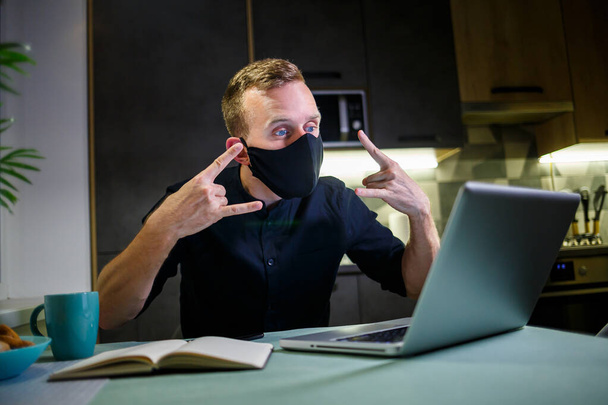Mladý obchodník v černé antivirové ochranné masce, pracuje doma, sedí u kuchyňského stolu s laptopem. Práce doma v karanténě - Fotografie, Obrázek