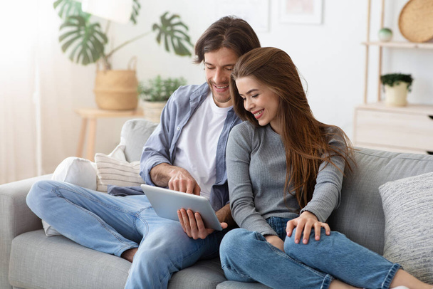 Man and woman sitting on sofa at home, using tablet - Фото, изображение