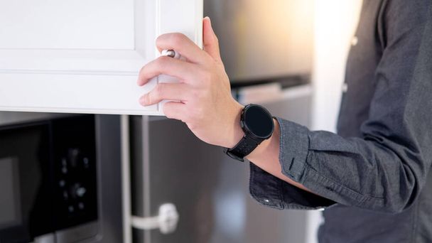 Αρσενικό χέρι άνοιγμα λευκό ντουλάπι πόρτα κοντά στο ψυγείο και φούρνο μικροκυμάτων στην οικιακή κουζίνα. Αρχική έννοια βελτίωσης επίπλων - Φωτογραφία, εικόνα