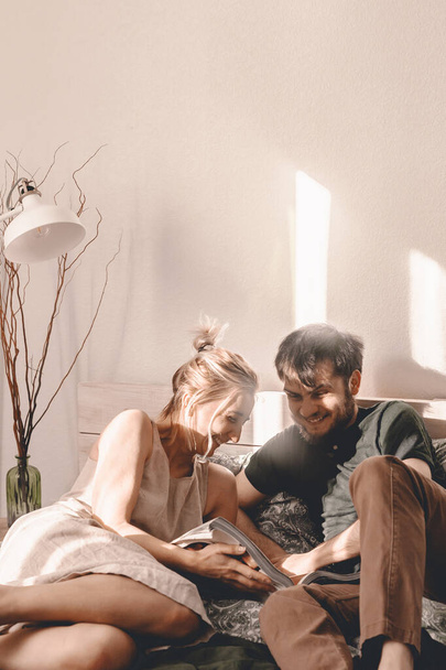 Happy couple is spending time in bed. Weekend leisure time. Time together. Stay home. Relax in bed. Modern bedroom. Read, eat, laugh. Lying in bed together. Bright modern bedroom. Direct sunlight. - Photo, Image