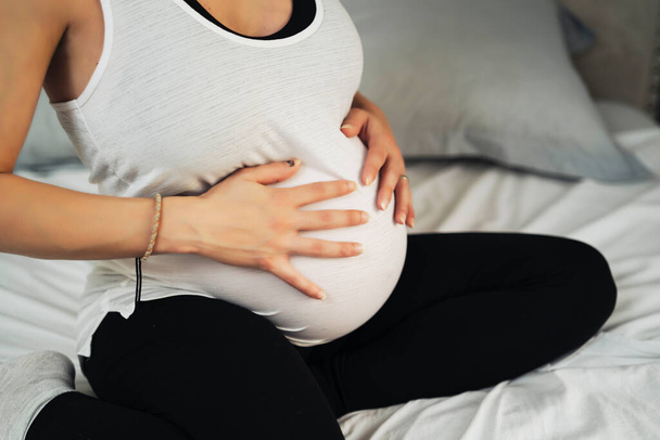 Gesneden beeld van mooie zwangere vrouw aanraken van haar buik zitten op het bed in haar kamer - Foto, afbeelding
