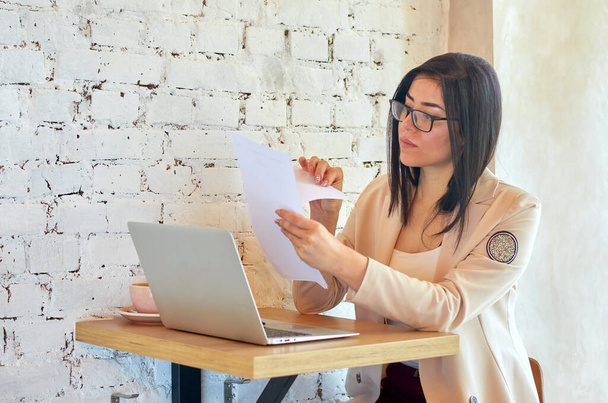 Jonge serieuze zakenvrouw met bril werkt in een trendy café met een witte laptop. Meisje in een licht jasje en rode broek. - Foto, afbeelding