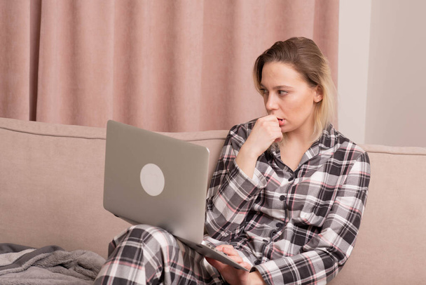 Kaunis nuori tyttö istuu sohvalla pyjamassaan ja on huolissaan siitä, mitä hän näki kannettavassa tietokoneessa.
. - Valokuva, kuva