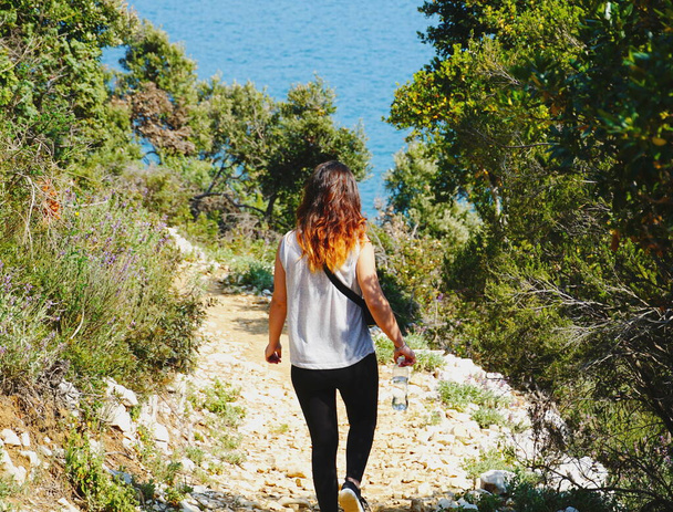 Woman hiking alone to pathway in the picturesque nature in a green forest by the sea - Photo, Image