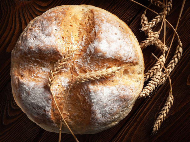 Homemade round bread. Top view of a loaf of warm fresh peasant bread and wheat spikelets on a wooden background. Homemade baking.  - Photo, Image