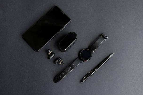 Accessories for men's beauty on a monochromatic background. black pen, black smart watch, smartphone and wireless headphones on dark background. View from above. Minimalist black trend 2020. - Photo, Image