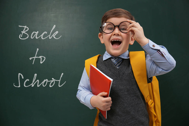 Cute little child wearing glasses near chalkboard with phrase BACK TO SCHOOL - Foto, Imagem