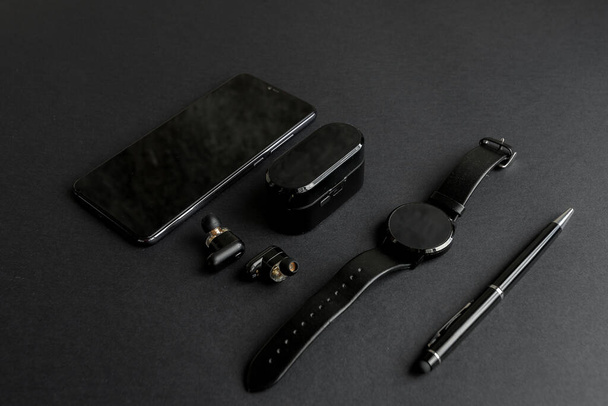 Accessories for men's beauty on a monochromatic background. black pen, black smart watch, smartphone and wireless headphones on dark background. View from above. Minimalist black trend 2020. - Фото, зображення