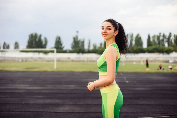 Красивая кудрявая девушка, напрягающая мышцы на стадионе. Девушка занимается спортом. Светло-зеленый костюм. Горизонтальное фото
. - Фото, изображение