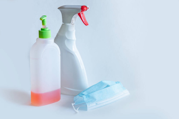 The bottle spray of white color with the red button, a liquid soap and medical mask is on a table of the white color, a set during a pandemic for disinfection, protection of respiratory system - Fotó, kép