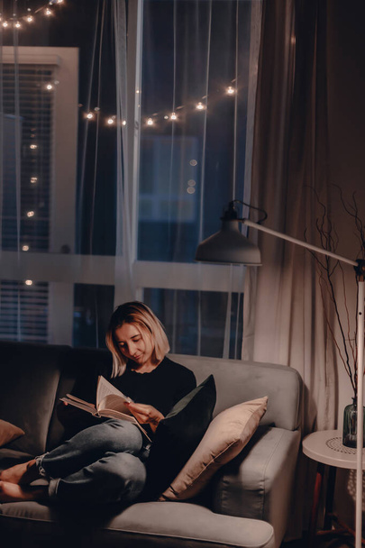 Girl reads book in the evening on couch. Night lamp light. Relaxing evening. Large window. Modern living room. Velvet pillows. Night light. Blonde woman reads fiction book. Quarantine stay at home. - Foto, Imagen