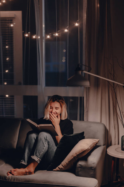Girl reads book in the evening on couch. Night lamp light. Relaxing evening. Large window. Modern living room. Velvet pillows. Night light. Blonde woman reads fiction book. Quarantine stay at home. - Zdjęcie, obraz