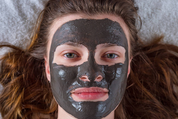 Młoda kobieta z włosami i maską kosmetyczną z czarnej gliny na twarzy - zabiegi kosmetyczne zbliżenie - Zdjęcie, obraz