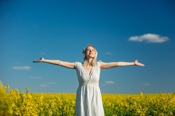 Beautiful woman in dress with headphones in rapeseed field in spring time - Photo, Image