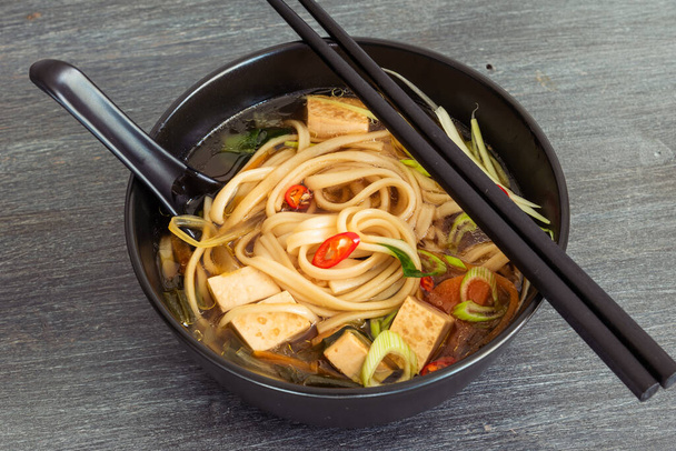 Японский веганский суп с удонной лапшой, тофу и овощами в черной миске с ложкой и палочками, вид сверху, крупный план
 - Фото, изображение