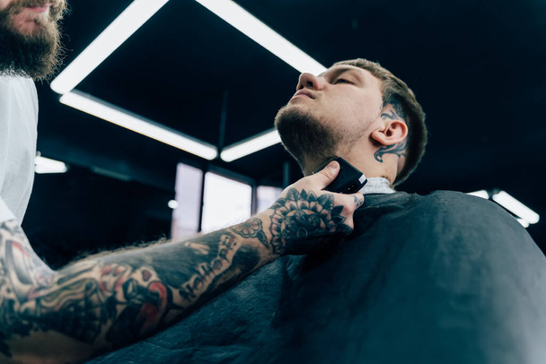 Tattooed κουρέας κουρέματος γενειοφόρος άνθρωπος με μηχανή ξυρίσματος στο κουρείο. Διαδικασία κομμωτικής. Κοντινό πλάνο ενός κομμωτή που κόβει τα γένια ενός γενειοφόρου αρσενικού. - Φωτογραφία, εικόνα