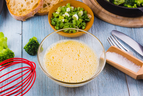 Bata ovos em uma tigela e ingredientes para fazer uma omelete com brócolis
 - Foto, Imagem