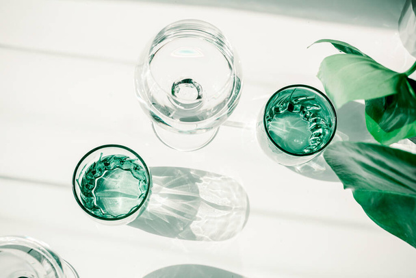 Ποτήρια με νερό στο λευκό τραπέζι. Σκληρό φως μέσα από γυαλιά, σκιές. Καθαρή μινιμαλιστική σύγχρονη τέχνη. Εννοιολογική εικόνα. Πράσινα και λευκά καθαρά χρώματα. Καθαρό νερό σε γυαλί. Απλή οπτική έννοια. - Φωτογραφία, εικόνα