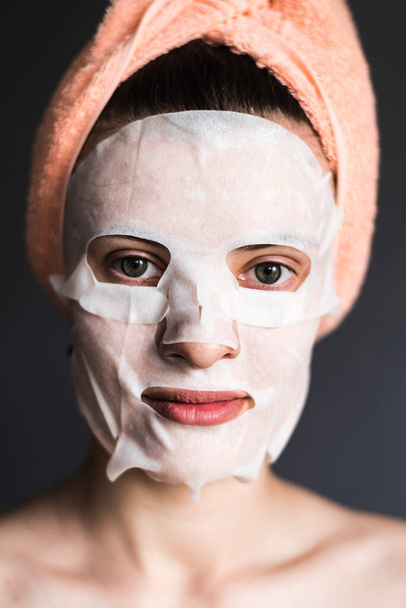 Mujer joven con máscara de lámina cosmética sobre fondo oscuro - concepto de cuidado facial
 - Foto, imagen