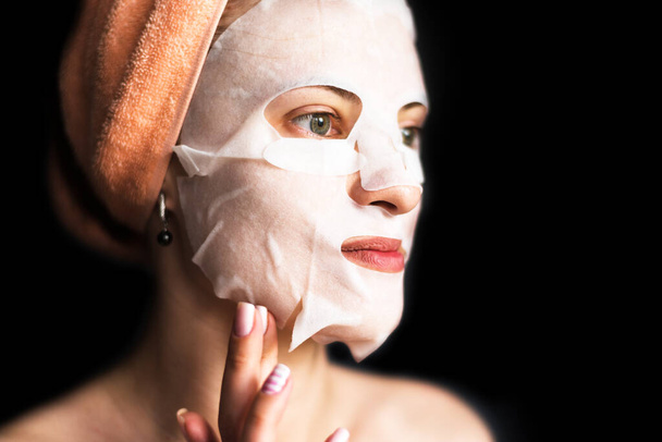 Mujer joven con máscara de lámina cosmética sobre fondo oscuro - concepto de cuidado facial
 - Foto, imagen