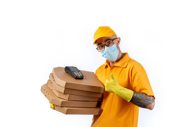 A cautious young man from a courier delivery service in a medical mask and gloves holds boxes with pizza and a terminal for cashless payment on a white background - Фото, зображення