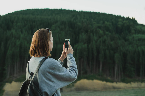 Hiker girl in casual clothes and with rbkzak takes pictures of natural mountain scenery with coniferous forest on smartphone camera, view from back. Hiker woman makes photo of mountain on smarphone - Фото, зображення