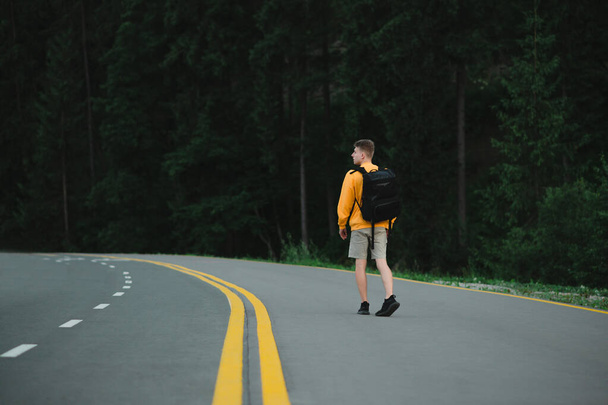Νεαρός τουρίστας σε casual ρούχα και σακίδιο με τα πόδια κατά μήκος ενός δασικού δρόμου και κοιτάζοντας γύρω με ένα σοβαρό πρόσωπο. Κομψός τύπος που περπατάει σε ασφαλτοστρωμένο δρόμο.. - Φωτογραφία, εικόνα