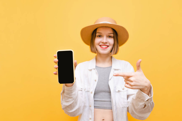 Happy lady holds a black screen smartphone in her hand and points at it with her finger, looks into the camera and smiles, wears a hat and light clothes. Copy space - Фото, изображение