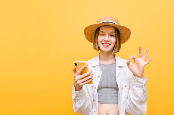 Açık renk elbiseli ve şapkalı güzel bir kızın portresi elinde akıllı telefonla kameraya bakıyor ve sarı arka planda izole edilmiş güzel bir jest gösteriyor. Boşluğu kopyala - Fotoğraf, Görsel