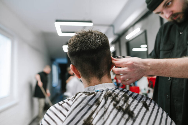 Homme coiffeur coupe les cheveux du client avec tondeuse à la main, sourit sur fond de mur blanc, se concentrer sur les mains du maître. Serious barber crée une coiffure à la mode pour le client
. - Photo, image