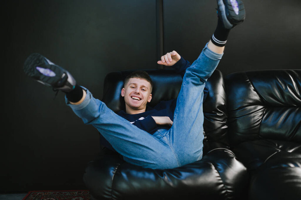 Pozitív fiatalember alkalmi ruhában fekve egy fekete bőrkanapén feltartott lábakkal, a kamerába nézve és mosolyogva. Vidám srác szórakozás kanapén sötét fal háttér. - Fotó, kép