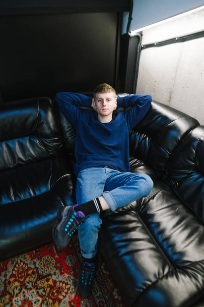 Attractive young man in casual clothes and stylish sneakers resting on a black leather sofa in a dark room, looking into the camera with a serious face. Vertical. - Photo, Image