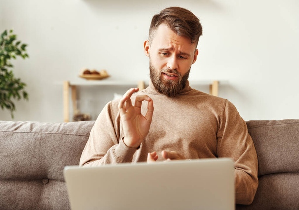 Serious young man in casual clothes working from home during coronavirus talking to colleague and showing okay gesture while having online conversation and discussing work issue - Photo, Image