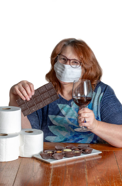Maskeli bir kadın evde kalıp çikolata, şarap ve tuvalet kağıdıyla yemek yemeye hazır. - Fotoğraf, Görsel