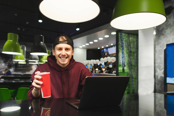 Portrait happy guy in cap and burgundy hoodie sitting in fast food restaurant with laptop and glass of cold cola drink, looking at camera and smiling.Student drinks a cool drink and uses the internet - Photo, Image