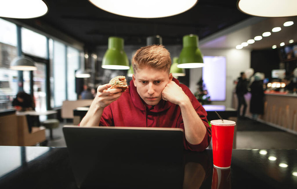 Un joven serio se sienta en una mesa de café, tiene una hamburguesa apetitosa en sus manos y mira la pantalla de un portátil. Estudiante enfocado trabaja en una cafetería y come comida rápida
. - Foto, imagen