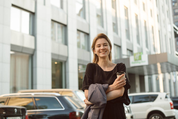 Mujer alegre con amplia sonrisa sosteniendo café y chaqueta caminando cerca del edificio de oficinas y estacionamiento. Sonriente jovencita sintiéndose feliz paseando por la calle urbana
. - Foto, Imagen