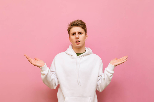 Surprised guy stands on a pink background with his hands in the sides, staring confusedly at the camera with a serious face. Puzzled guys in white hoodie isolated on pink background. - Photo, Image