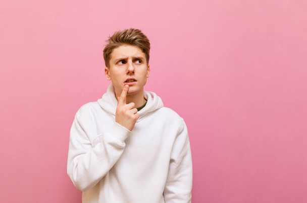 Thoughtful guy in white roe deer stands on a pink background and looks away at copy space with a serious face. Serious young man with blond hair looks away and thinks. Isolated. - 写真・画像